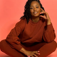 Load image into Gallery viewer, Black smiling girl in cinnamon colored jogger set sitting down in front of a pink background.
