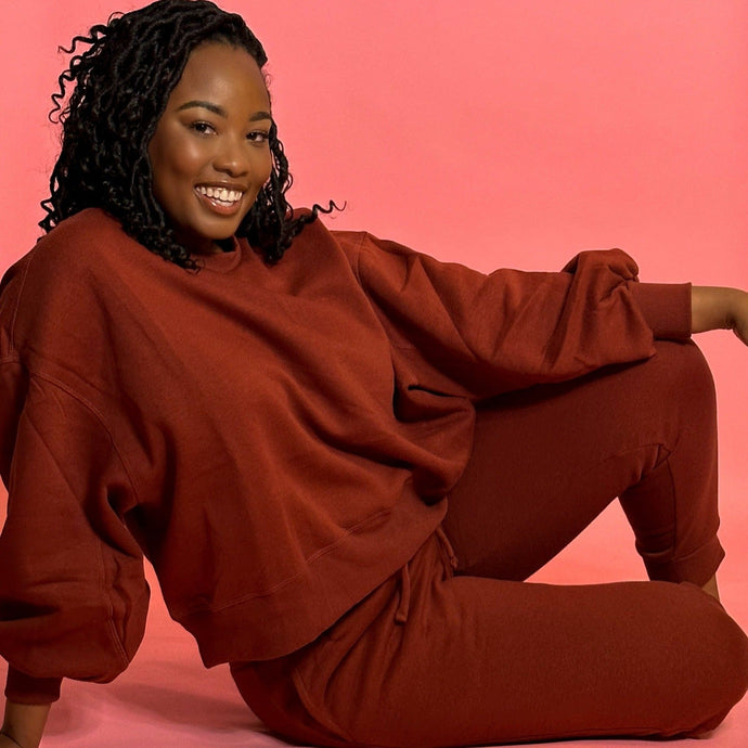 Black smiling girl in cinnamon colored jogger set sitting down in front of a pink background.