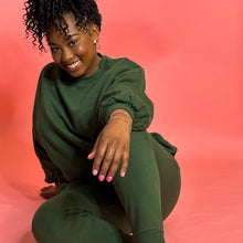 Load image into Gallery viewer, Black girl smiling and wearing green joggers set and sitting down in front of pink background
