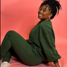Load image into Gallery viewer, Black girl smiling and sitting on pink background in green jogger set.
