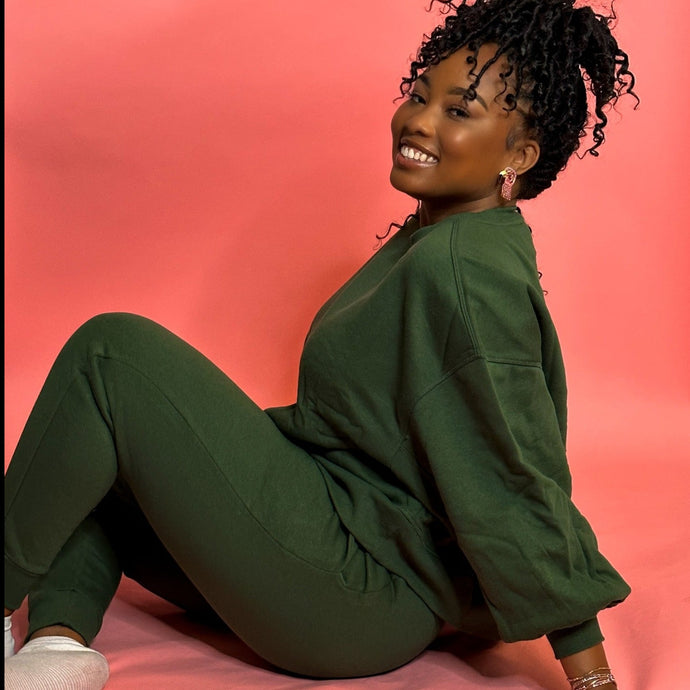 Black girl smiling and sitting on pink background in green jogger set.
